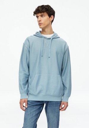 Jeune homme portant un sweat à capuche bleu clair avec cordons et poche avant, associé à un jean bleu, regardant légèrement vers la gauche.
