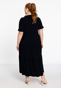 Dunkelblaues, gestuftes Maxikleid mit kurzen Rüschensäumen, einem Kragen und sanft fallendem Stoff. Getragen mit weißen Riemchenschuhen.