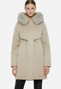 Cappotto imbottito beige con un ampio colletto in pelliccia grigia, tasche laterali e vita svasata. Il design presenta una linea dritta e una cerniera frontale.