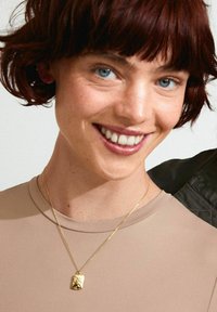 Goldkette mit einem kleinen, strukturierten Anhänger, getragen über einem beigen Oberteil. Der Anhänger ähnelt einer kleinen Tasche mit eckigen Details.