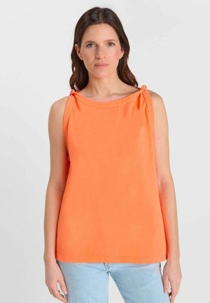 Mujer con cabello castaño, vistiendo una camiseta sin mangas naranja con tirantes atados en los hombros y pantalones vaqueros azul claro, de pie contra un fondo blanco liso.