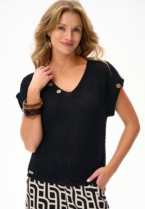 Kobieta w czarnym dzianinowym topie z krótkim rękawem z drewnianymi guzikami i geometrycznej spódnicy w czarno-białe wzory, pozująca z ręką przy twarzy.