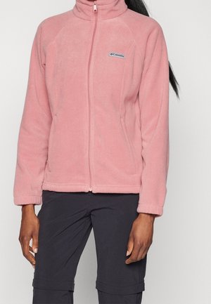 Osoba ubrana w jasnoróżową, zapinaną na zamek błyskawiczny polarową kurtkę z logo Columbia i ciemne spodnie, stojąca na jednolitym jasnym tle.