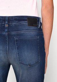 Vista trasera de una persona vestida con jeans de mezclilla azul oscuro con detalles descoloridos y un parche de marca negro, combinados con una blusa rosa clara.