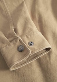 Beige stofærme med en manchet med knapper, med metalsnapknapper prydet med mærkenavnet "GABBA".