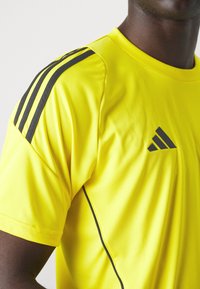 T-shirt de sport jaune avec des rayures noires sur les épaules et un logo. Fabriqué en matériau respirant, avec un col rond et des manches courtes.