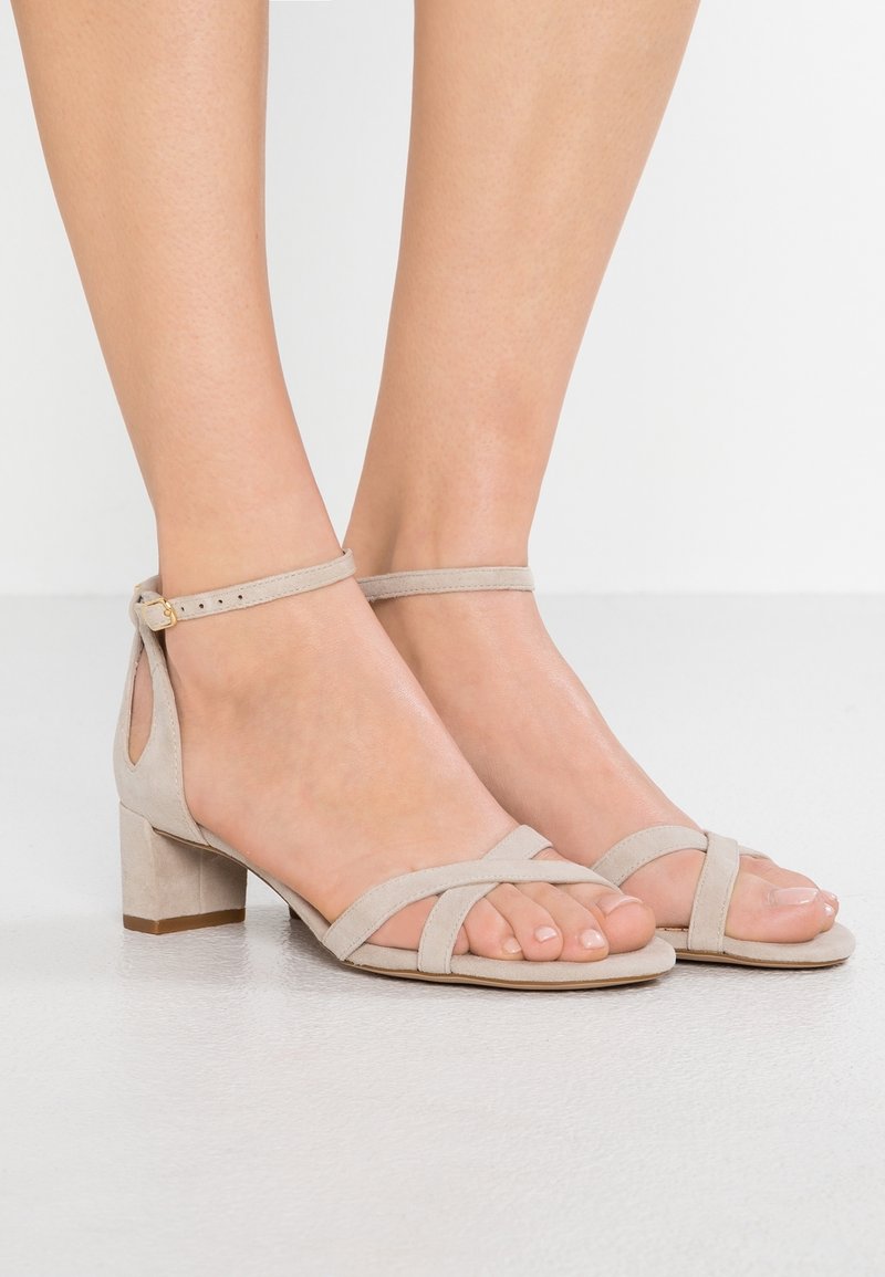 Nohy obuté v béžových semišových sandálech na blokovém podpatku s řemínky kolem kotníků a otevřenou špičkou na pozadí bílé podlahy a zdi.