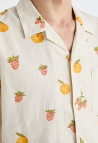 Camisa de lino de manga corta, base crema con patrones de naranja y fresa, que presenta un cuello con muescas y cierre de botones.