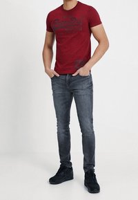 Hombre vistiendo una camiseta gráfica roja ajustada, jeans oscuros ajustados y zapatillas negras, de pie con las manos en los bolsillos contra un fondo liso.