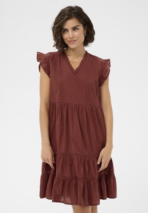 Femme aux cheveux courts et bouclés portant une robe sans manches rouge rouille avec des épaules volantées et une jupe à volants, debout devant un fond uni.