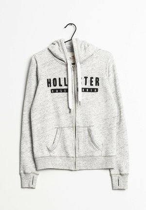 Veste à capuche zippée gris clair avec poches avant, cordons blancs, et "Hollister California" imprimé en noir sur la poitrine, suspendue à un cintre noir.