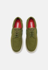 Green canvas shoes with a lace-up design, rounded toe, and contrasting red insoles. Features textured fabric and a subtle white rubber sole.