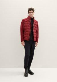 Czerwona pikowana kamizelka z wysokim kołnierzem, bocznymi kieszeniami i zapięciem na zamek, noszona na czarnym swetrze, w połączeniu z granatowymi spodniami i czarnymi botkami.