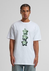 White cotton t-shirt featuring a vertical graphic of three green stars with a black outline, relaxed fit, round neckline, and short sleeves.