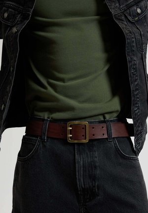 Mann trägt grünes Hemd, schwarze Jeansjacke, schwarze Jeans und braunen Ledergürtel mit quadratischer goldener Schnalle.