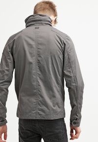 Szara, lekka kurtka z wysokim kołnierzem, teksturowaną tkaniną, widocznymi szwami i guzikami na mankietach; zaprojektowana na dopasowany krój.