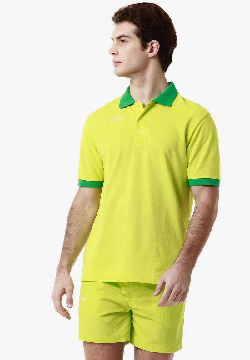 Polo shirt giallo neon con colletto e polsini verdi, caratterizzato da un piccolo logo bianco sul petto, abbinato a pantaloni corti coordinati.