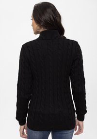 Sort kabelstrikke-sweater med høj hals og lange ærmer, der har teksturerede mønstre. Materialet ser blødt og kraftigt ud.