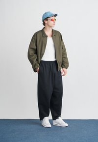 Alyvuogių žalios spalvos bomber stiliaus striukė, balti marškinėliai be rankovių, juodos laisvos kelnės, mėlyna kepurė, daugiaspalvės saulės akiniai ir balti sportbačiai. Paprasta fono.