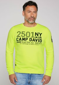 Sudadera de color amarillo neón con mangas largas, letras negras y escote redondo. Presenta un parche en la manga izquierda y puños acanalados.