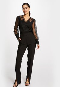 Haut noir avec des manches transparentes à pois et un décolleté en V bordé de dentelle, associé à un pantalon noir ajusté avec des fentes au niveau des chevilles.
