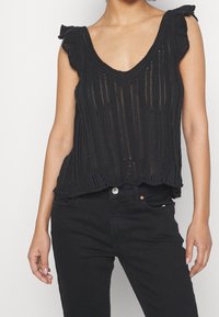 Svart stickad linne med V-ringning, med utsvängda ärmar, öppen vävstruktur och vågig nederkant, i kombination med svarta jeans.