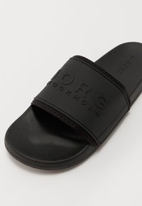 Black slide sandal with a textured footbed and a wide strap featuring embossed brand name. Made from rubber material, flexible design.