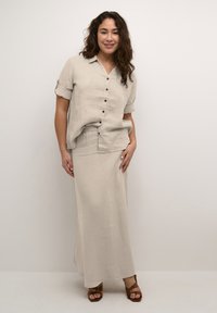 Vrouw met golvend haar die een beige shirt met knopen en opgerolde mouwen draagt, samen met een bijpassende lange rok, poseert blootsvoets tegen een neutrale achtergrond.
