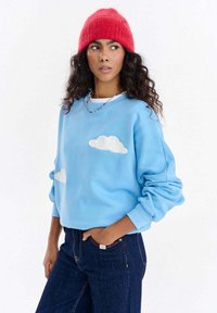 Jasnoniebieska bluza z haftem białych chmur na przodzie, zestawiona z ciemnymi dżinsami i czerwonym dzianym beanie.