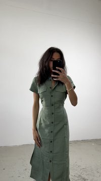 Zelena lanena haljina s kratkim rukavima, dugmadi sprijeda, dva džepa na prsima i bočnim prorezima. Glatka tekstura s prilagođenim krojem.