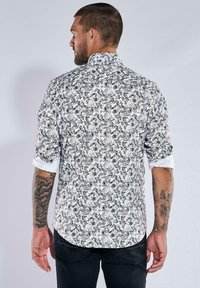 Weißes Hemd mit kompliziertem schwarzen Paisleymuster, lange Ärmel hochgekrempelt und hoher Kragen. Getragen mit dunklen Jeans, die detaillierte Tattoos zur Schau stellen.