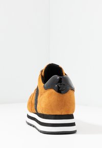 Zapatilla deportiva de ante en color naranja con detalles en charol negro. Presenta una suela gruesa de rayas blancas y negras y un cuello acolchado.