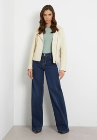 Giacca da moto in pelle crema sopra una blusa verde chiaro, abbinata a jeans in denim scuro a vita alta e gamba larga, con tacchi a punta.