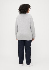 A person with curly hair wears a gray sweater and dark jeans, standing with their back to the camera against a plain white background.