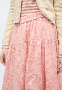 Γυναίκα που φοράει μπεζ πλεκτή ζακέτα πάνω από ροζ και λευκή ριγέ μπλούζα, συνδυασμένη με ροζ φούστα με υφή και λουλουδάτο μοτίβο.