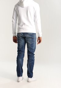 Sudadera blanca con capucha, jeans de denim azul con una forma ajustada, textura desgastada y detalles de costura visibles, combinados con zapatillas blancas.
