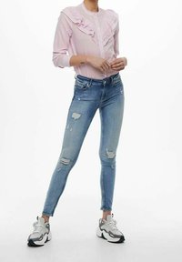 Camicia rosa con bottoni e volant, abbinata a jeans skinny blu con dettagli strappati e sneakers grigie con accenti bianchi.