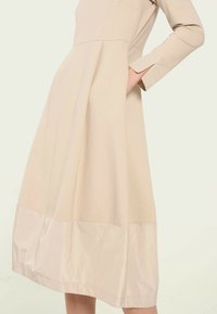 Rochie bej cu maneci lungi, cu un bust ajustat și o fustă evazată. Prezintă un tiv inferior din satin, buzunare laterale și un design simplu, curat.