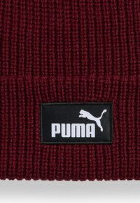 Bordeaux gebreide beanie met een geribbelde textuur. Bevat een zwart rechthoekig label met een wit Puma-logo en inscriptie.