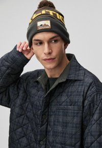Nawlekana granatowa kurtka z zielonym kołnierzem i kieszenią; zestawiona z brązową i żółtą czapką z pomponem oraz logo góry.