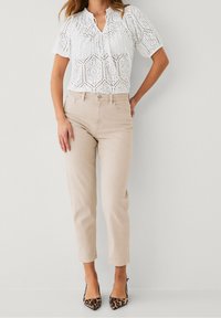 Femme portant un haut blanc à manches courtes en broderie anglaise, un jean beige taille haute et des escarpins à bout pointu en imprimé léopard, se tenant devant un fond blanc.