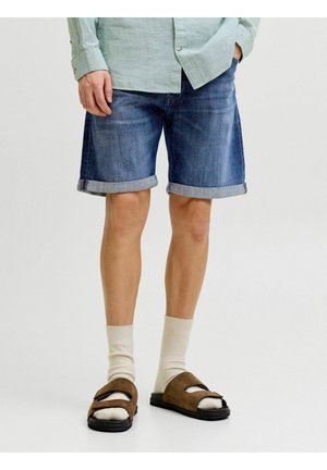 Mężczyzna noszący podwinięte dżinsowe szorty, jasnozieloną koszulę z długim rękawem, kremowe skarpetki i brązowe sandały z odkrytymi palcami, stojący na białym tle.