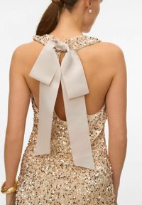 Mirdzoša bēša kleita ar atvērtu muguru, kurai ir fliteru apdare, sasietas satīna lentes bēšā tonī un gludi pieguļošs siluets.