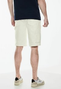 Pantalones cortos tipo cargo beige con dos bolsillos traseros y un bolsillo lateral; combinados con zapatillas blancas y grises con acentos negros.