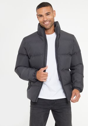 Dunkelgraue Daunenjacke mit hohem Kragen, elastischen Bündchen und Reißverschluss vorne. Getragen über einem weißen T-Shirt und schwarzen Jeans.