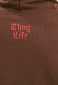 Felpa marrone con il testo rosso "Thug Life" in un font audace e stilizzato sul retro. Il materiale sembra liscio con una texture morbida.