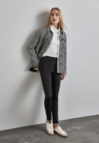 Veste en pied-de-poule noir et blanc, chemise blanche à boutons, leggings noirs, baskets blanches avec des accents beige, se tenant devant un fond gris.
