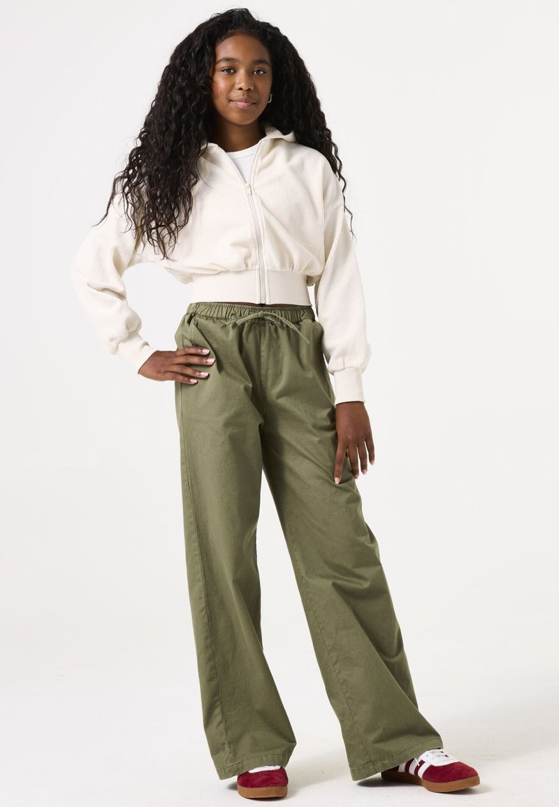 Veste cropped beige clair zippée, pantalon vert à jambes larges avec une taille élastique, et baskets rouges avec des rayures blanches.