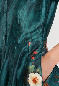 Teal satin dress featuring button front, floral embroidered accents in red and cream, with a flared silhouette and side pockets.