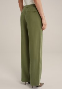 Pantalones de pierna ancha en verde oliva, con cintura elástica, textura suave y bolsillos traseros sutiles. Ajustados hacia el dobladillo, combinados con zapatos de punta.